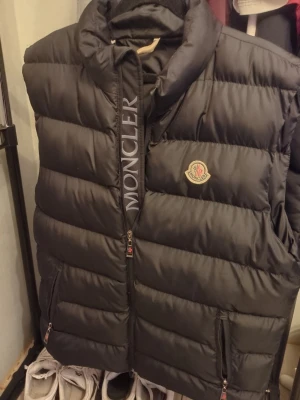 Svart dunväst från Moncler - Snygg svart dunväst från Moncler med hög krage och dragkedja framtill. Västen har Moncler-logga på bröstet och stora fickor med dragkedja. Puffig design och klassisk Moncler-etikett på insidan. Perfekt för lager-på-lager och streetwear.