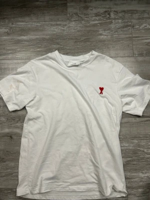 Vit AMI t-shirt med rött hjärta - Vit t-shirt i bomull med rund hals och korta ärmar. På bröstet finns en broderad röd hjärtlogga med ett A, vilket ger en stilren och trendig look. Perfekt för dig som gillar minimalistisk design med en twist.