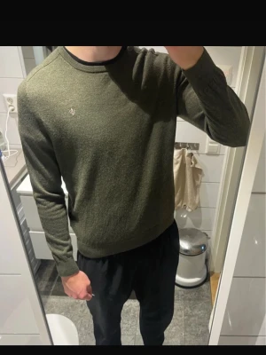 Mörkgrön stickad tröja Morris - 100% merino mörkgrön stickad tröja från Morris med rund halsringning och diskret broderad logga på bröstet. Tröjan har långa ärmar och ribbade muddar vid ärmslut och nederkant. Perfekt för lager-på-lager och stilrena outfits.
