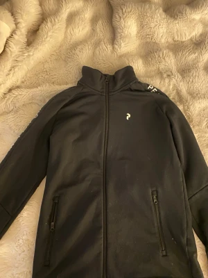 Svart ziphoodie från Peak Performance - Säljer en svart hoodie från Peak Performance med dragkedja framtill och två fickor med dragkedja. Logga på bröstet och texten 'Peak Performance' längs båda ärmarna. 