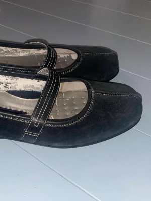 Svarta ballerinaskor med rem - Stilrena, sköna svarta ballerinaskor i mockaimitation med bred rem över foten och vita kontrastsömmar. Skorna har rund tå och låg sula, vilket ger en modern och trendig look. Perfekta för dig som gillar klassiska men ändå unika detaljer.