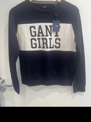 Mörkblå stickad tröja GANT GIRLS - Mörkblå stickad tröja från GANT med vit panel över bröstet och texten 'GANT GIRLS' i svart. Klassisk rund halsringning och långa ärmar. Perfekt för dig som gillar stilren och sportig look.