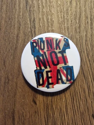 Punks Not Dead pinsknapp - Cool knapp med texten 'Punks Not Dead' i svart graffiti-stil. 4,4 cm. Perfekt accessoar för dig som gillar punk och vill sticka ut. Rund form och klassisk punkkänsla.