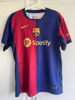 FC Barcelona matchtröja blå/röd - Säljer en officiell FC Barcelona matchtröja från Nike i klassiska blå och röda färger. Tröjan har klubbmärke, Nike-logga och Spotify som sponsor framtill samt UNHCR-logga bak. Korta ärmar och sportig passform, perfekt för fotbollsträning eller support.