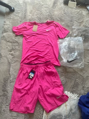 Rosa träningsset från Nike - Snyggt rosa träningsset från Nike med t-shirt och shorts. Båda plaggen har Nike-logga i vitt och är gjorda i ett lätt och snabbtorkande material, perfekt för träning eller sport. T-shirten har rund hals och shortsen har resår i midjan med dragsko.