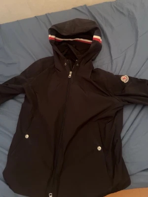 Svart vindjacka med huva Från moncler - Snygg svart vindjacka med huva och dragkedja framtill. Jackan har två fickor med silverfärgade knappar och Moncler-logga på ärmen. Huvan har röd, vit och blå randig kant. Perfekt för blåsiga dagar.