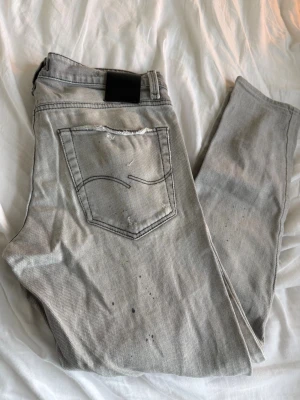 Jack & Jones jeans - Säljer ett par grå jeans från Jack & Jones med slitningar och distressed detaljer på både fram- och baksida. Jeansen har fem fickor, normal midja och raka ben. Perfekta för en avslappnad och trendig look.