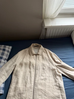 Beige linnéovershirt med dragkedja - Stilren beige overshirt i linnematerial med dragkedja framtill och en bröstficka. Klassisk krage och raka ärmar ger en avslappnad look. Perfekt lager för vår och sommar, lätt att matcha med olika outfits.