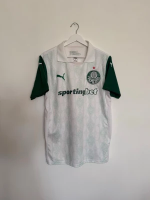 Vit Palmeiras matchtröja från Puma - Snygg vit matchtröja från Palmeiras med gröna ärmar och detaljer. Tröjan har klubbmärke och Puma-logga på bröstet samt sponsortryck 'sportingbet' på framsidan. Tillverkad i lätt och ventilerande material, perfekt för fotboll eller träning.