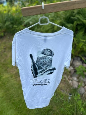 Notre Succés T-shirt - Säljer en vit t-shirt med tryck på ryggen som föreställer en stad, champagneflaska och klocka samt texten 'Dolce Vita Portofino'. Framtill står det 'Yacht Club Notre Succès'. T-shirten har rund hals och korta ärmar. Perfekt för dig som gillar stilrena plagg med coola detaljer.