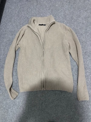 Beige ribbad ziptröja från 157 - Säljer en beige ribbstickad ziptröja från 157 med hög krage och dragkedja framtill. Tröjan har långa ärmar och är gjord i ett mjukt, stickat material som passar perfekt för lager på lager. Enkel och stilren design som funkar till det mesta.