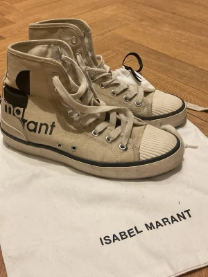 Beiga Isabel Marant high top sneakers - Snygga high top sneakers från Isabel Marant i beige canvas med svart logga på sidan och vit gummisula. Skorna har klassisk snörning, svarta detaljer och ribbad tå. Perfekta för en trendig och avslappnad stil.