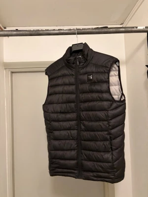 Svart dunväst från Lindbergh - Snygg svart dunväst från Lindbergh med quiltad design och hög krage. Västen har dragkedja framtill och en diskret logotyp på bröstet. Perfekt att slänga över en hoodie eller tröja för extra värme. Materialet är lätt och smidigt. En liten defekt (se bild 4) som man inte tänker på när man bär västen.  Nypris 1499. Kan gå ner i pris vid en snabb affär!