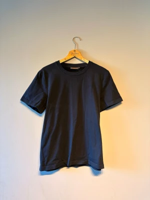 Mörkblå basic t-shirt med broderi - En stilren mörkblå t-shirt i bomull med korta ärmar och rund hals. På bröstet finns ett diskret broderat emblem som ger en clean look. Perfekt till jeans eller shorts för en enkel och snygg outfit.