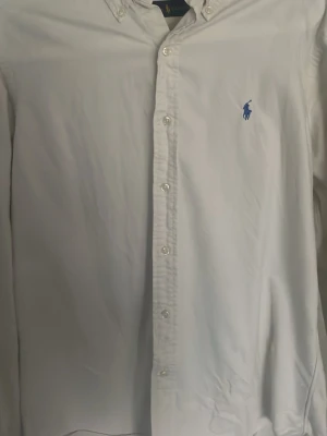 Vit skjorta från Ralph Lauren - Klassisk vit skjorta från Ralph Lauren med blå broderad logga på bröstet. Skjortan har knappar hela vägen fram och button-down krage. Tillverkad i bomull och har lång ärm. Perfekt för en clean och stilren look.