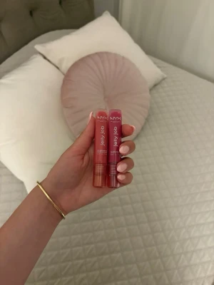 NYX Jelly Bab läppglans duo - Två NYX Jelly Bab läppglans i tub, en i persikorosa och en i bärig rosa nyans. Båda har genomskinliga plastförpackningar med vita detaljer och praktisk applikator. Perfekta för att ge glans och färg till läpparna.