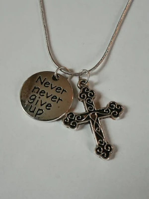 Halsband med kors och textberlock - Snyggt halsband i silverfärg med två hängen: ett runt med texten 'Never never give up' och ett dekorativt kors med detaljerade mönster. Kedjan är tunn och smidig. Materialet rostfritt stål 