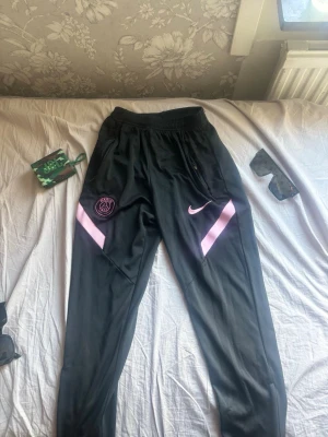 Svarta PSG träningsbyxor Nike - Säljer ett par svarta träningsbyxor från Nike med PSG-logga på ena benet och rosa detaljer. Byxorna har dragkedjor vid fickorna och är tillverkade i ett lätt, sportigt material som passar perfekt till träning eller chill.
