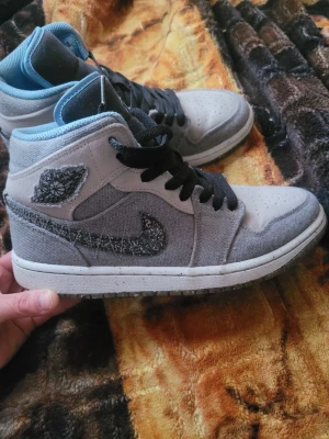 Gråa höga sneakers med svart swoosh - Säljer ett par gråa höga Air Mid crator Jordan sneakers med svart broderad swoosh och svarta skosnören. Skorna har blå detaljer vid ankeln och en vit sula. Ovandelen är i 100% mocca och textilmaterial och har perforeringar på tån för ventilation. Endast testad 
