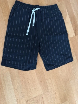 Svarta randiga linne shorts med snörning - Snygga svarta shorts med vita vertikala ränder och vit snörning i midjan. Shortsen har avslappnad passform och sidofickor, perfekta för sommardagar. Tillverkade i ett mjukt och lätt material som känns skönt mot huden.