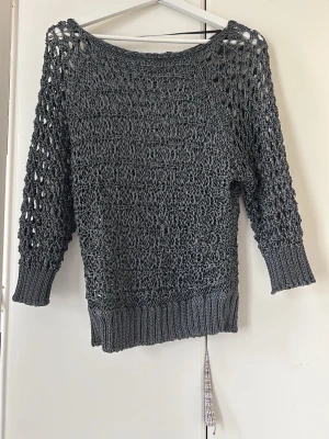 Mörkgrå stickad meshtröja - Säljer en mörkgrå stickad tröja med meshstruktur och trekvartsärmar. Tröjan har bred ribbstickad mudd vid ärmslut och nederkant samt en avslappnad passform. Perfekt att bära över ett linne eller t-shirt för en cool lager-på-lager-look.