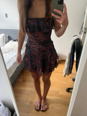 Mörk kort klänning med volang - Säljer en kort klänning i mörka toner med volangkant och figurnära passform. Klänningen har ett abstrakt mönster i svart och vinrött, är ärmlös. Storleken är 36 (S) men skulle säga att den även passar XS❣️