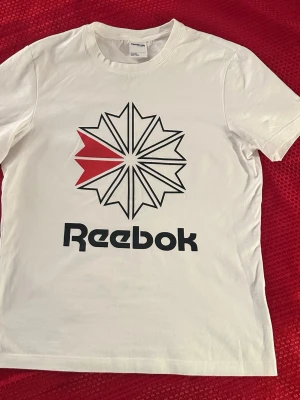 Vit Reebok t-shirt med stor logga S, cotton 100% - Säljer en vit t-shirt från Reebok med stor grafisk logga i svart och rött framtill. Klassisk passform och rund halsringning. Perfekt för en sportig och avslappnad stil.