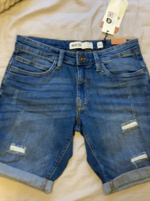 Blå jeansshorts från INDICODE - Säljer ett par blå jeansshorts från INDICODE med slitna detaljer och uppvikta benslut. Klassisk femficksmodell i bomull med knappgylf och normal passform. Perfekta för sommaren och riktigt snygga till en enkel t-shirt eller hoodie.