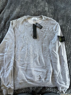Grå sweatshirt från Stone Island - Snygg grå sweatshirt från Stone Island med klassisk rund hals och ribbade muddar. Tröjan har den ikoniska Stone Island-loggan på vänster ärm och är tillverkad i mjuk bomull. Perfekt för en avslappnad och stilren look.