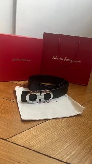 Svart skärp från Salvatore Ferragamo  - Snyggt svart skärp från Salvatore Ferragamo med en ikonisk silverfärgad dubbel-Gancio-spänne. Skärpet är tillverkat i slätt skinn och har en stilren, klassisk design som passar till många olika outfits.