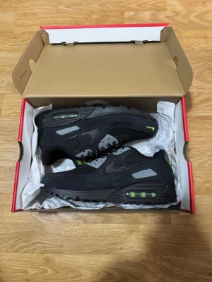 Nike Air Max 90 svarta sneakers - Nike Air Max 90 sneakers i svart med detaljer i grått och neongrönt. Skorna har en klassisk siluett med synlig Air Max-dämpning i sulan och är tillverkade i en mix av mocka och mesh. Swoosh-logga på sidan och svarta skosnören.
