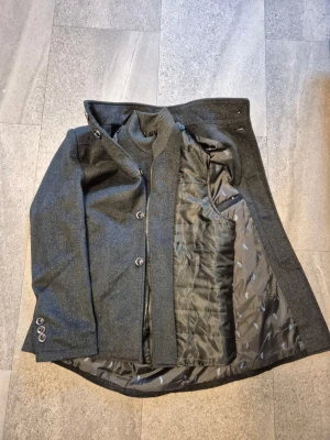 Grå ullkappa med kvalitet och stilren look! - Snygg grå kappa i ullmix med krage och dragkedja samt knappar framtill. Två fickor på sidorna och diskreta detaljer vid ärmsluten. Fungerar lika bra att ha på hösten som vintern. Figursydd med siden inuti så den sitter perfekt! Jackan som fungerar för allt.