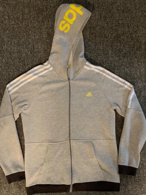 Grå adidas hoodie med dragkedja - Snygg grå hoodie från adidas med hel dragkedja, vita ränder på axlarna och gul adidas-logga på bröstet och huvan. Mjuk insida i fleece och svarta muddar vid ärmslut och nederkant. Perfekt för en sportig och avslappnad stil. Sitter som storlek M