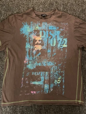 Brun t-shirt med tryck från Santino - Cool brun t-shirt från Santino med färgglatt tryck i blått, gult och rosa framtill. T-shirten har korta ärmar och kontrastsömmar längs sidorna. Perfekt för dig som gillar streetstyle och unika prints.