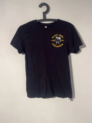 Svart Black Army Stockholm t-shirt - Svart t-shirt med tryck fram och bak. På bröstet står det 'Black Army Stockholm' med en figur och årtal i gult, och på ryggen står det 'A.C.A.B.' i stora gula bokstäver. Klassisk passform och rund hals.