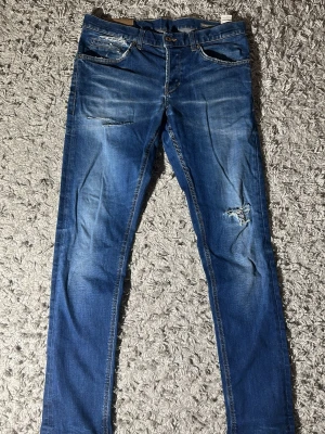 Blå slim fit dondup jeans med slitningar - Säljer ett par blå slim fit jeans med coola slitningar och detaljer på benen. Jeansen har klassiska fem fickor och en modern passform som sitter snyggt. Perfekt för dig som gillar en avslappnad och trendig look. Längd: 103 cm midja: 42cm