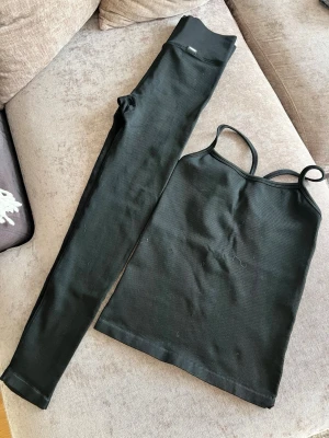 Svart ribbat  träningsset Stronger - Säljer ett svart ribbat set med linne och tights. Linnet har smala axelband och en rak passform, medan tightsen är höga i midjan och åtsittande. Materialet är stretchigt och mjukt. Storlek passar S M L. Köpt för 1400kr