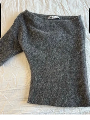 Grå fluffig stickad tröja från Zara - Mysig grå stickad tröja från Zara med fluffig och mjuk textur. Tröjan har en enkel design med långa ärmar och rund halsringning. Perfekt för att hålla sig varm under kyliga dagar. Materialet känns lyxigt 