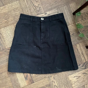 Svart kortkjol från Monki - Säljer en svart kortkjol från Monki med två stora fickor framtill och knappstängning i midjan. Den är lite dammig men annars i bra stick! Är i jeans liknande tyg men är väldigt mjuk