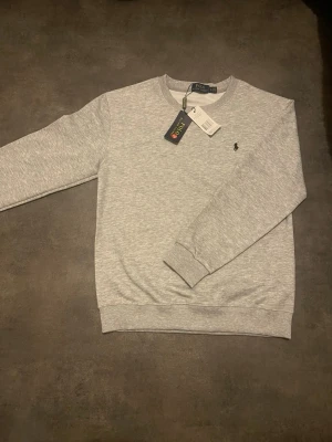 Grå sweatshirt från Polo Ralph Lauren - Klassisk ljusgrå sweatshirt från Polo Ralph Lauren med rund halsringning och diskret broderad logga på bröstet. Tillverkad i mjuk bomullsblandning och har ribbade muddar vid ärmslut och nederkant. Perfekt för en clean och avslappnad stil.