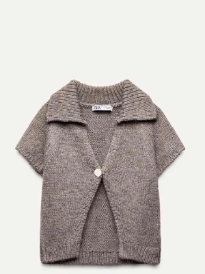 Kortärmad stickad kofta från Zara - Kortärmad stickad kofta i gråbeige ton från Zara. Koftan har en bred krage, ribbade detaljer vid ärmslut och nederkant samt en dekorativ knapp framtill. Perfekt för lager på lager och en avslappnad men trendig look.