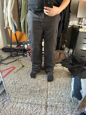 Mörkgrå cargobyxor med fickor - Säljer ett par mörkgrå cargobyxor med raka ben och klassiska sidofickor på låren. Byxorna har en avslappnad passform och är perfekta för en streetwear-look. Materialet känns slitstarkt och bekvämt, och de har bälteshällor i midjan.