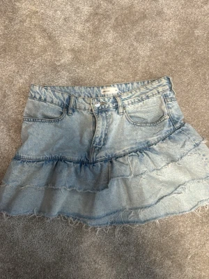 Volangig jeanskjol från Gina tricot - Säljer en ljusblå jeanskjol från Gina  med volangdetaljer och råa kanter. Kjolen har klassiska fickor fram men inga bak. Trendig och säljs inte längre 