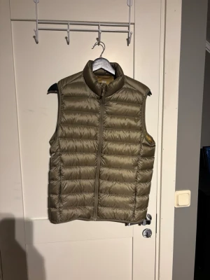 Olivgrön dunväst med hög krage - Snygg olivgrön dunväst med quiltad design och hög krage. Västen har dragkedja framtill och är lätt vadderad för extra värme. Perfekt att slänga över en hoodie eller tröja för en casual look. Passar dig som gillar stilrena och funktionella plagg.