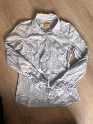 Ljusblå skjorta från ICHI - Klassisk ljusblå skjorta från ICHI med lång ärm och knappar framtill. Skjortan har en enkel krage och är gjord i mjuk bomull, perfekt för en clean och stilren look. Passar till jeans eller kjol för en avslappnad vibe.