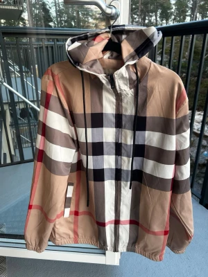 Beige vindjacka med rutigt mönster från burberry - Snygg beige vindjacka med klassiskt rutigt mönster i svart, vitt, rött och brunt. Jackan har huva med dragsko, dragkedja framtill och elastiska muddar. Perfekt för dig som gillar streetstyle och vill sticka ut med en trendig look.