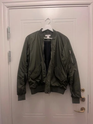 Olivgrön bomberjacka från H&M - Snygg olivgrön bomberjacka från H&M med klassisk passform. Jackan har ribbade muddar, två stora fickor framtill och en ärmficka med dragkedja. Tillverkad i ett glansigt material som ger en cool streetwear-look. Perfekt för dig som vill ha en stilren och trendig jacka.