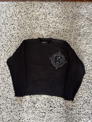 Svart stickad tröja från Vicinity - Svart stickad tröja från Vicinity med ett stort, vitt broderat logomärke på bröstet. Tröjan har rund halsringning och ribbstickade muddar vid ärmslut och nederkant. Perfekt för dig som gillar streetwear och vill ha en unik detalj i garderoben.