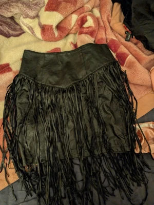 Svart skinnkjol med fransar - Cool svart kjol i skinn med långa fransar runt om som ger en edgy och unik look. Kjolen har en hög midja och kort passform, perfekt för att sticka ut. Passar dig som gillar statement-plagg och vill ha något extra till garderoben.