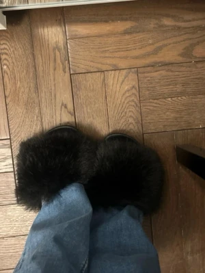Svarta fluffiga tofflor - Säljer ett par svarta tofflor med breda, fluffiga band i fuskpäls. Sulan är platt och gjord i syntetmaterial, perfekt för att glida runt hemma med en extra cozy vibe. Tofflorna har en modern och trendig look som passar till chill outfits.